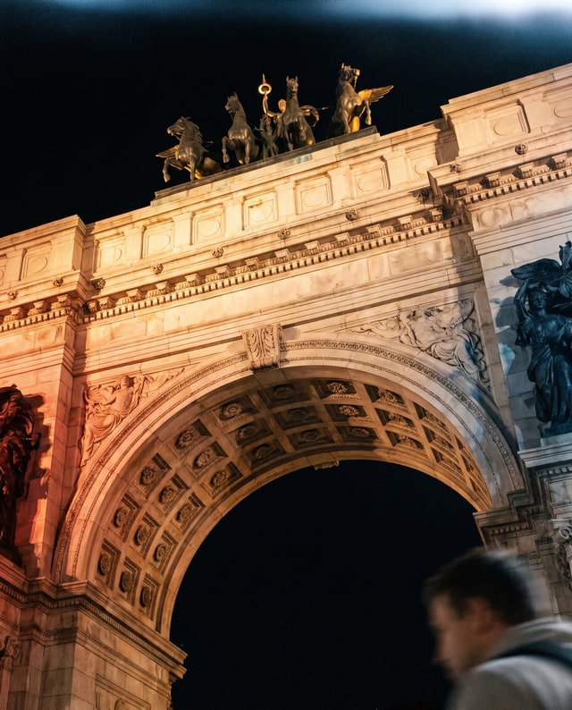 A warmly lit triumphal arch with a passerby in the foreground, made by Muse.