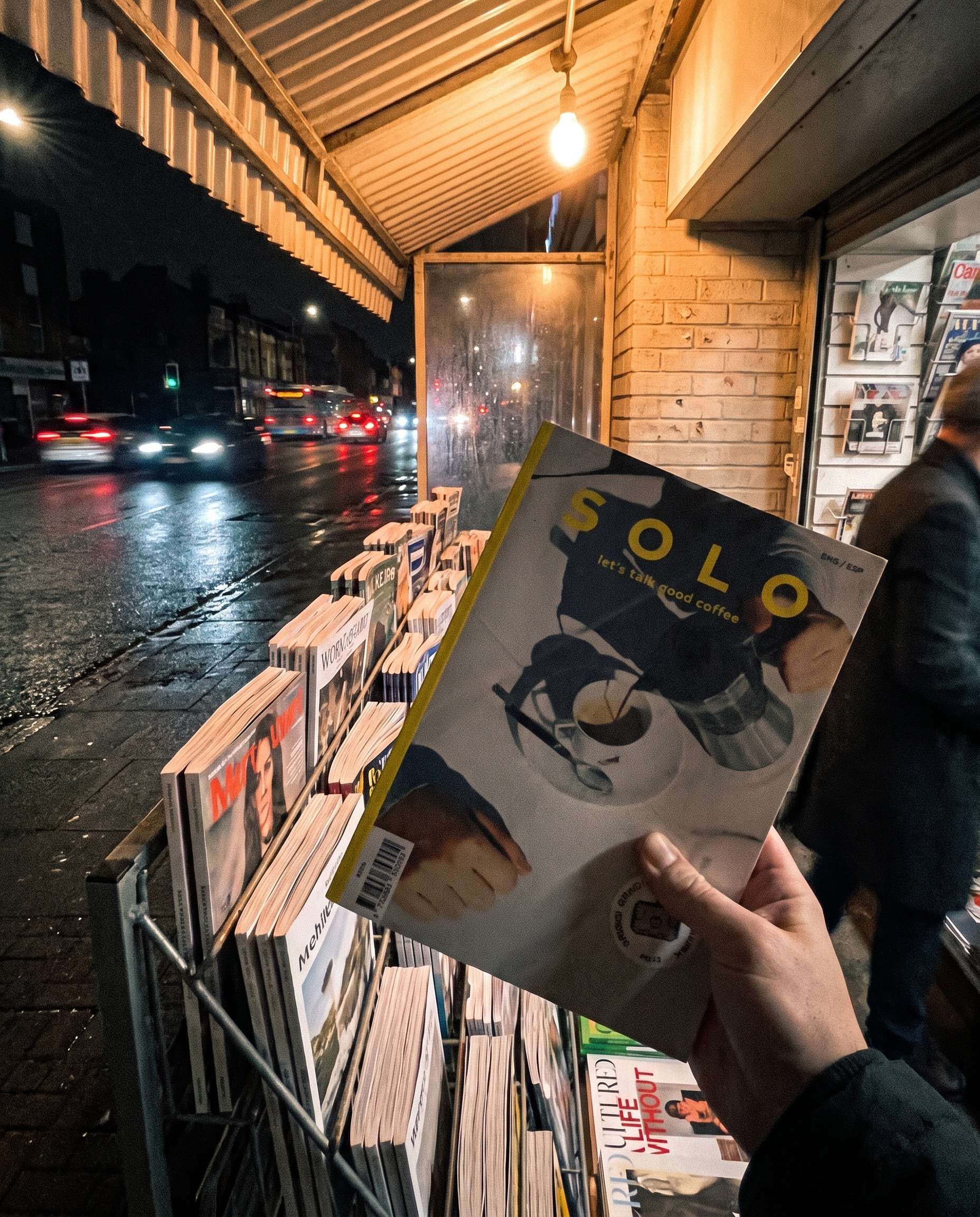 A hand holding a magazine outside a newsstand on a rainy night, made by Muse.