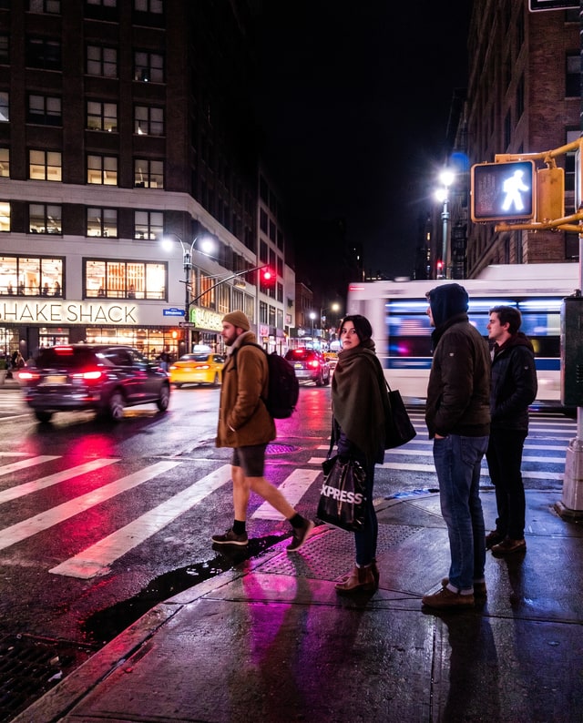 Pedestrians waiting at a wet city crossing under neon lights, made by Muse.